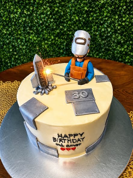 Welder-themed birthday cake with a handmade figure in a welding helmet, silver metal details, and a glowing spark